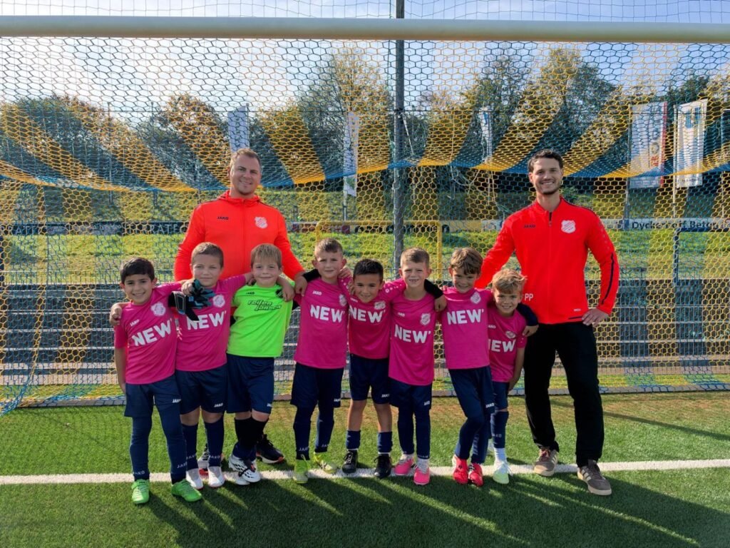 F-Junioren in pinken Trikots mit ihren beiden Trainern in orangen Jacken vor dem Tor