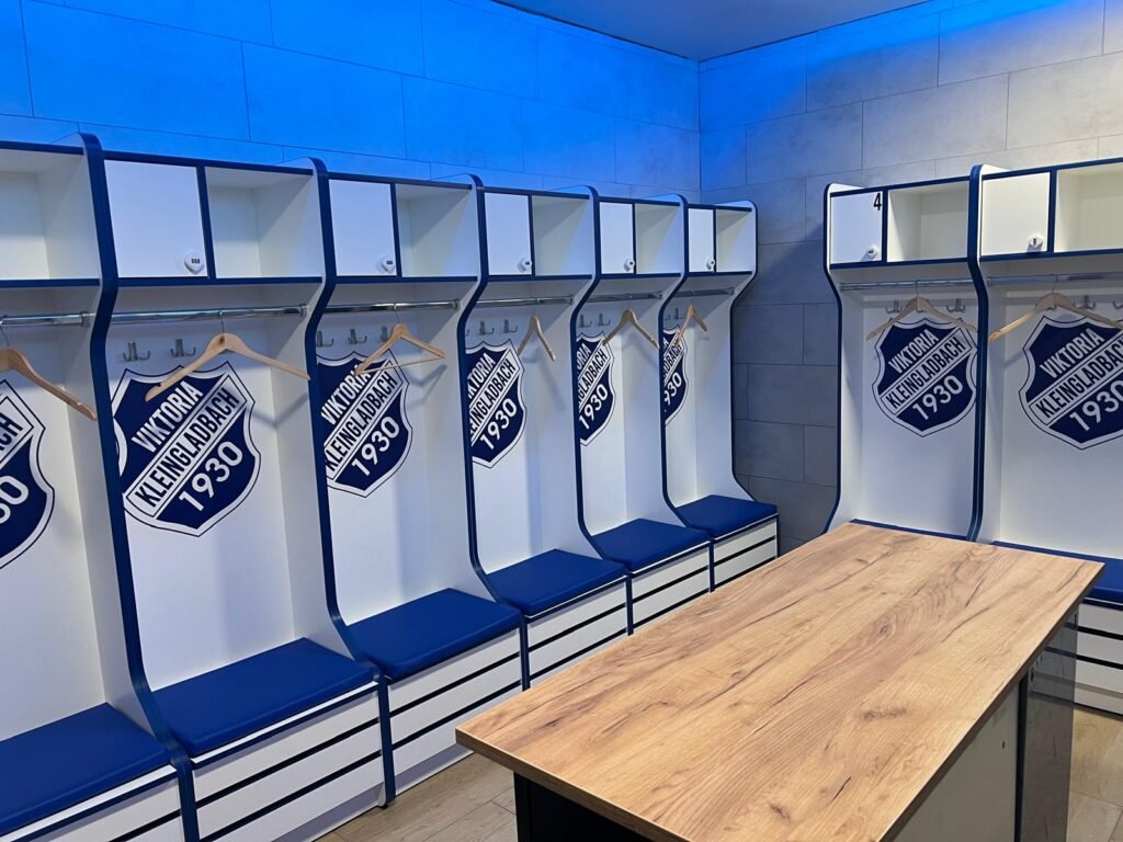 Neue Heimkabine mit weißen Spinden, Vereinswappen und blau gepolsterten Sitzbänken