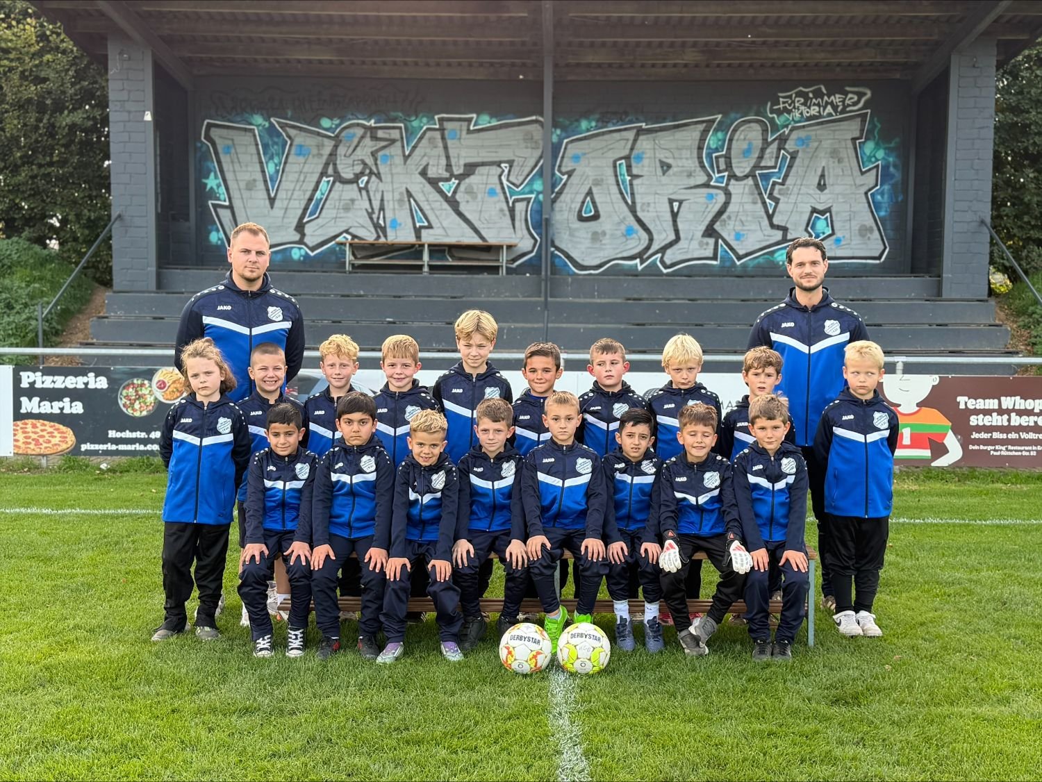 Teamfoto der F-Junioren des SV Viktoria Kleingladbach mit ihren Trainern vor der Tribüne mit Viktoria-Graffiti